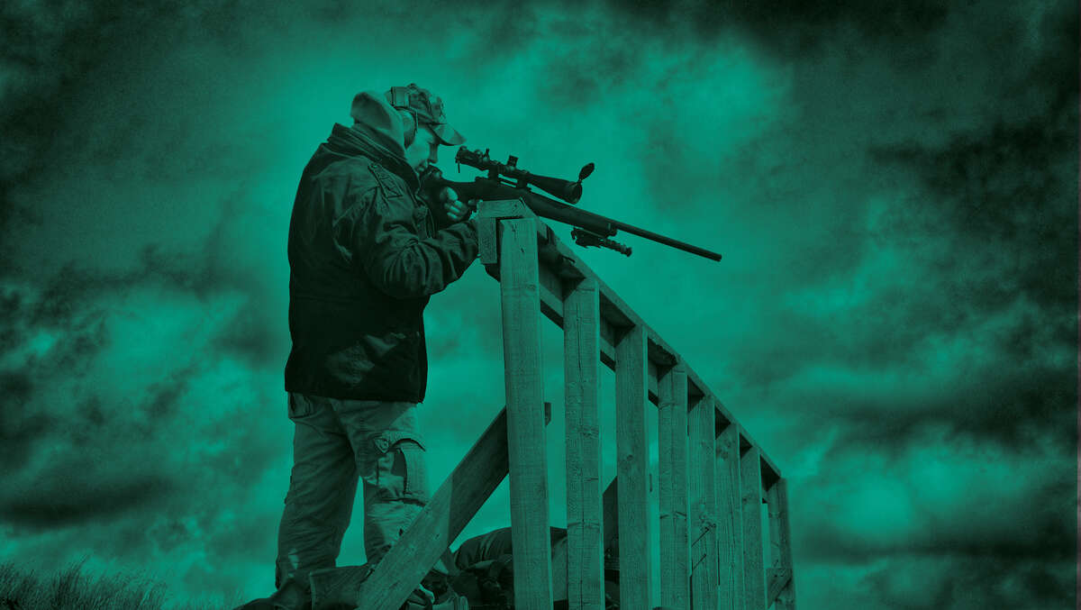 A man with a rifle using a fence post as a rest.