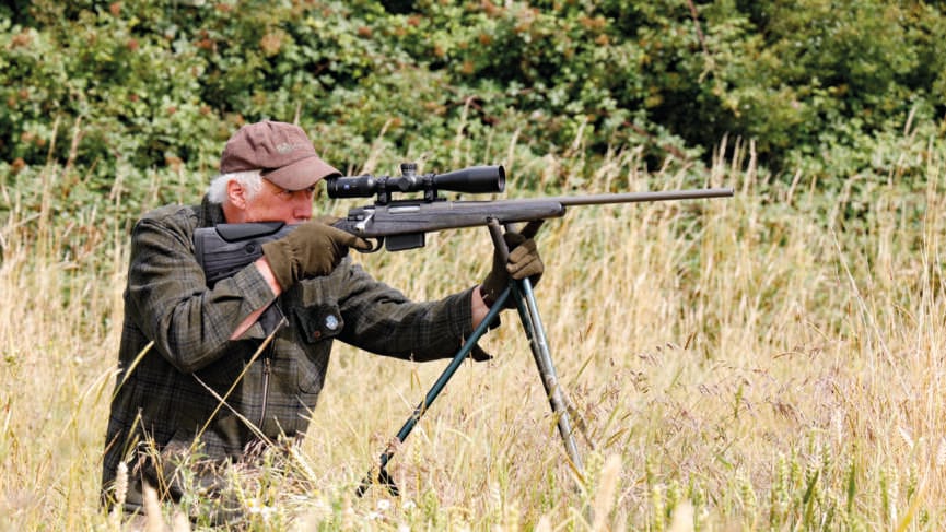 man in field aiming rifle through a scope on a tripod