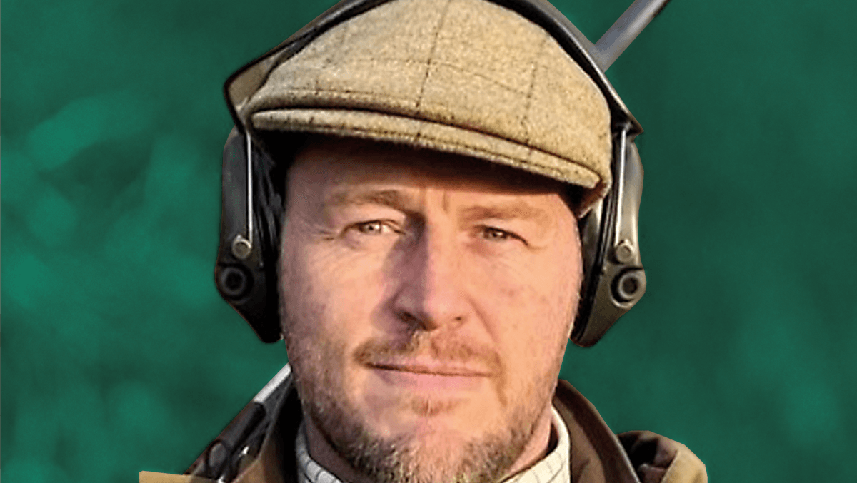 man wearing ear protectors and a flat cap holding a rifle